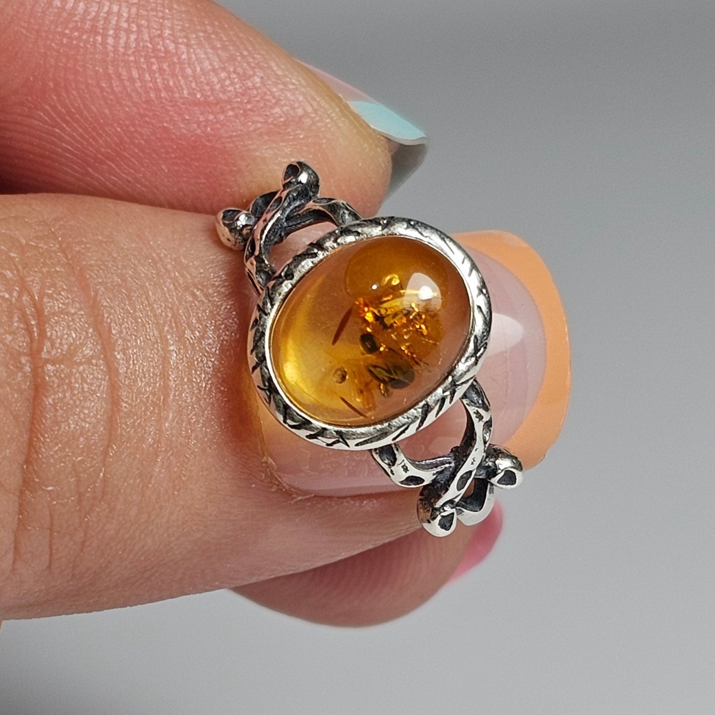 This adjustable ring is crafted from sterling silver and features a beautiful oval shaped light Amber stone with a textured bezel and criss-cross designed shoulders.