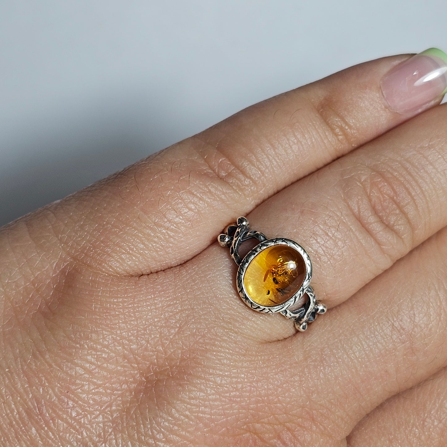 This adjustable ring is crafted from sterling silver and features a beautiful oval shaped light Amber stone with a textured bezel and criss-cross designed shoulders.