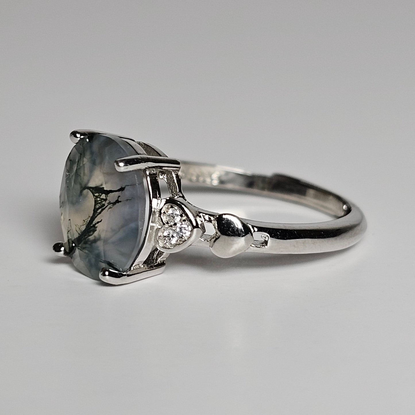 This adjustable ring is crafted from sterling silver and features a stunning oval shaped Moss Agate stone with zircon and heart designed shoulders.