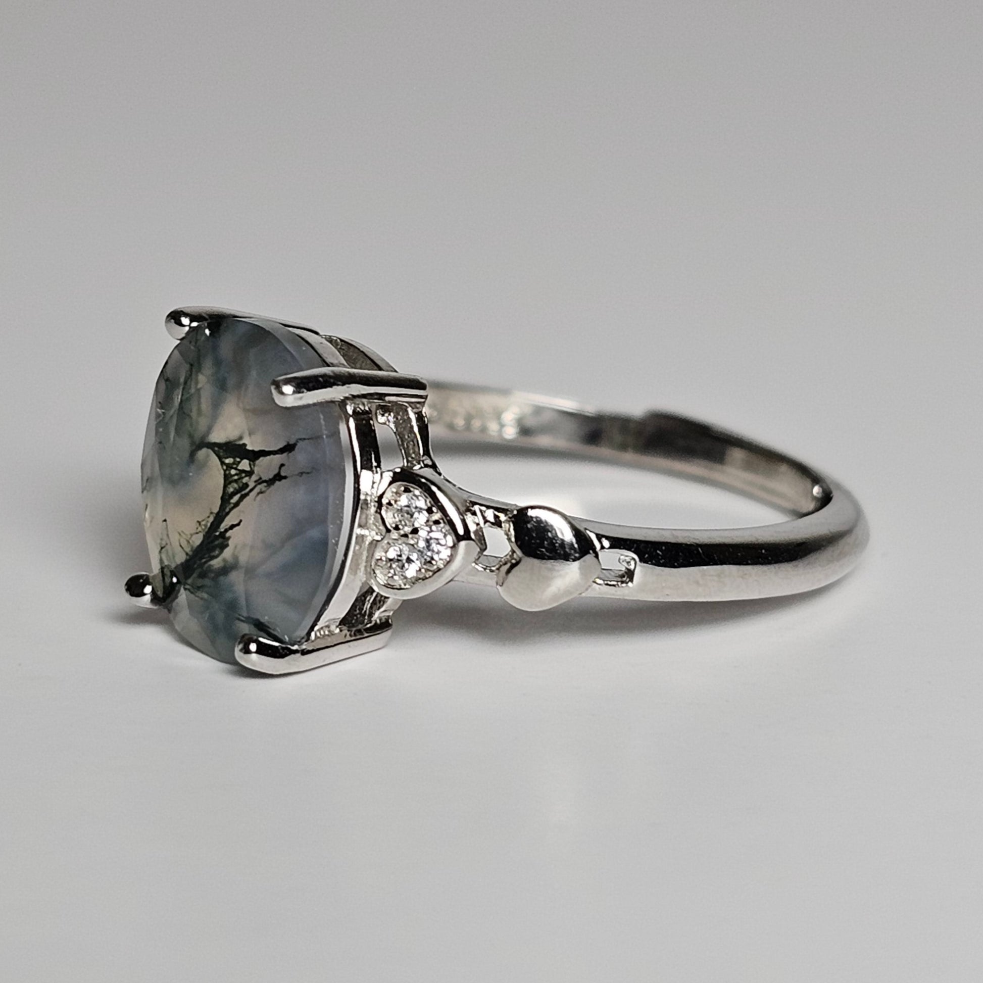 This adjustable ring is crafted from sterling silver and features a stunning oval shaped Moss Agate stone with zircon and heart designed shoulders.