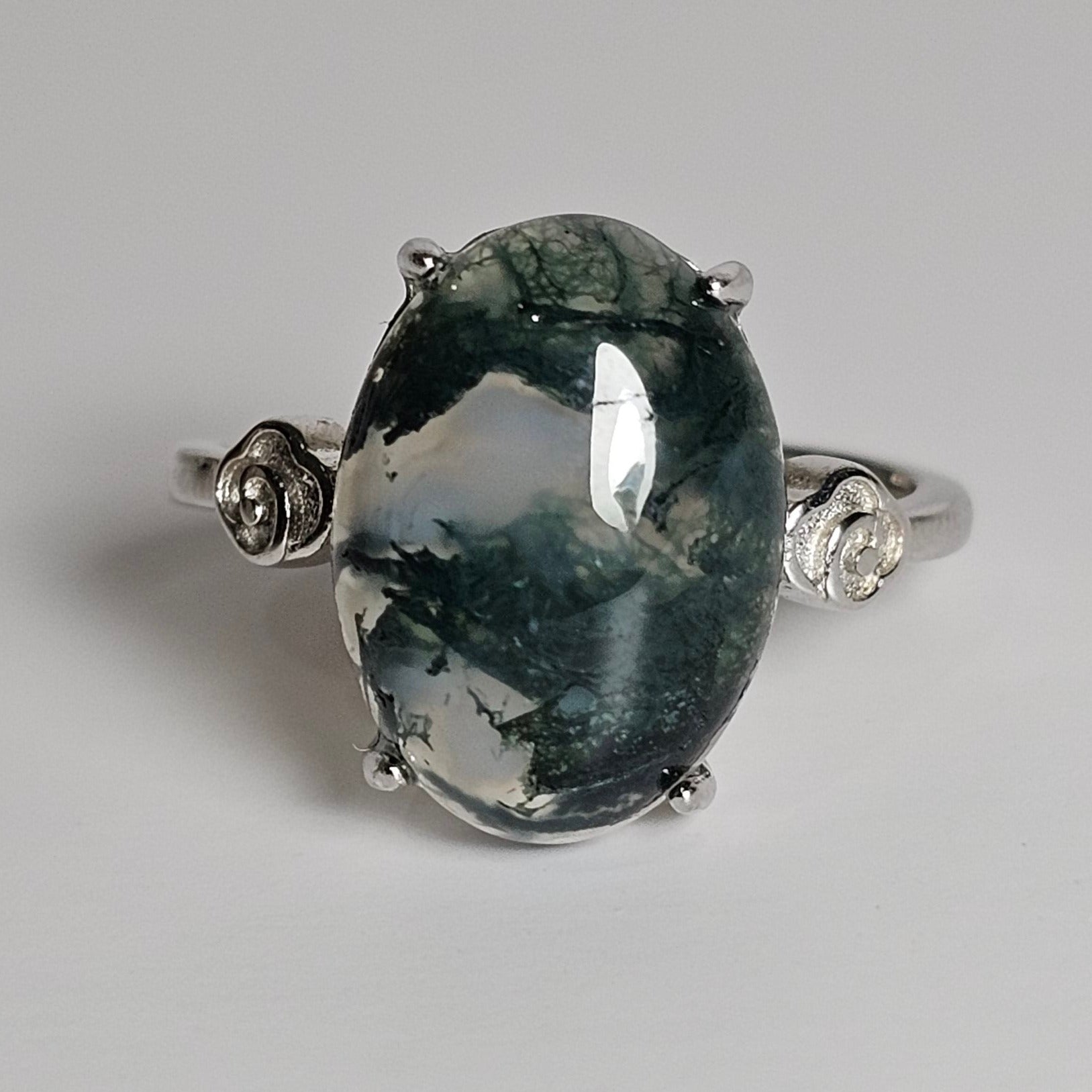 This adjustable ring is crafted from sterling silver and features a stunning oval shaped Moss Agate with rose details on the shoulder of the ring band.