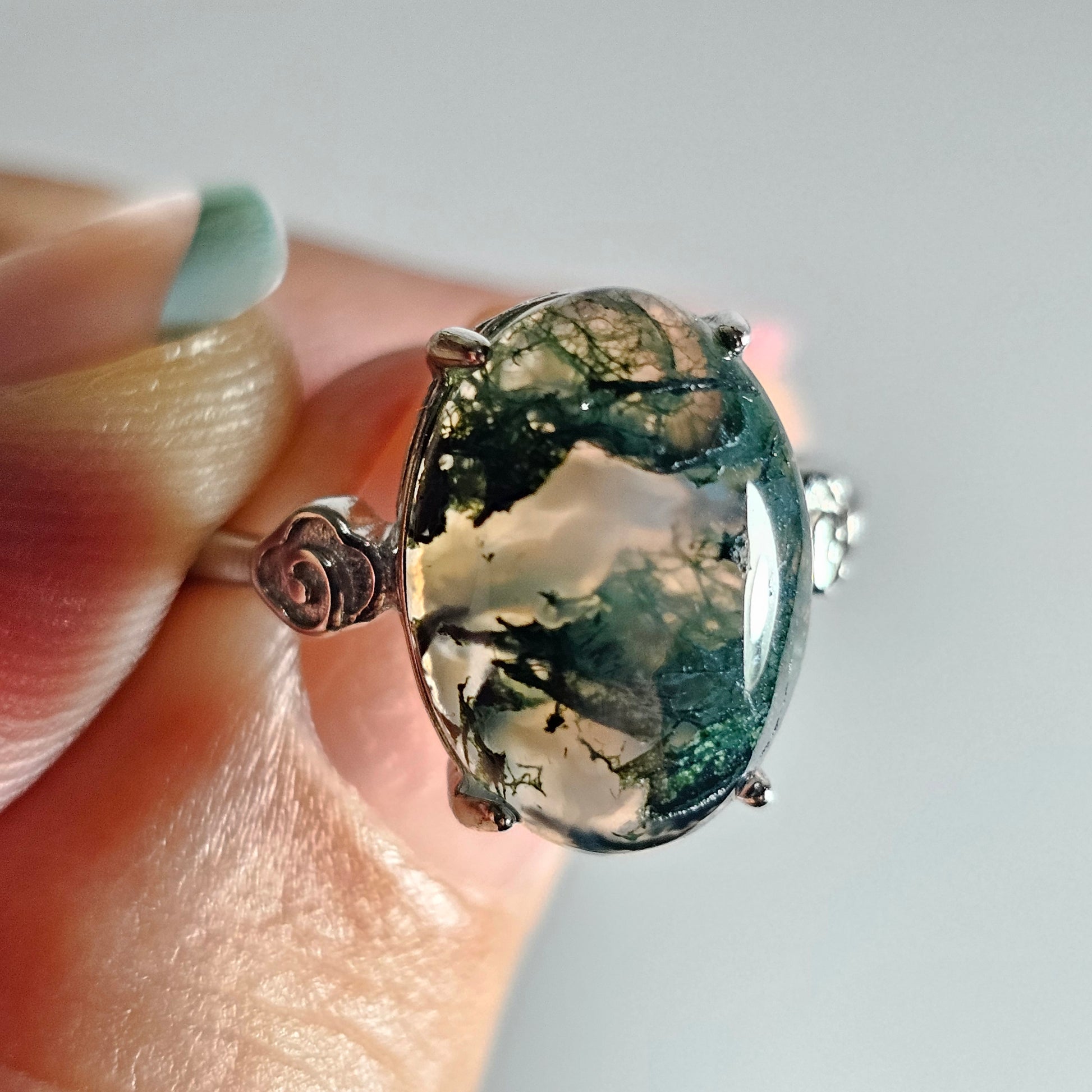 This adjustable ring is crafted from sterling silver and features a stunning oval shaped Moss Agate with rose details on the shoulder of the ring band.