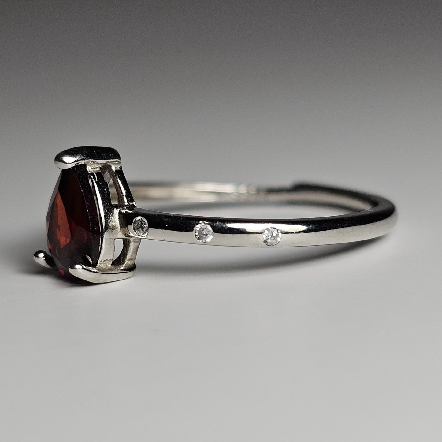 This adjustable fine ring is crafted from sterling silver and features a pear cut Garnet with zircon inlays along the shoulders of the ring band.