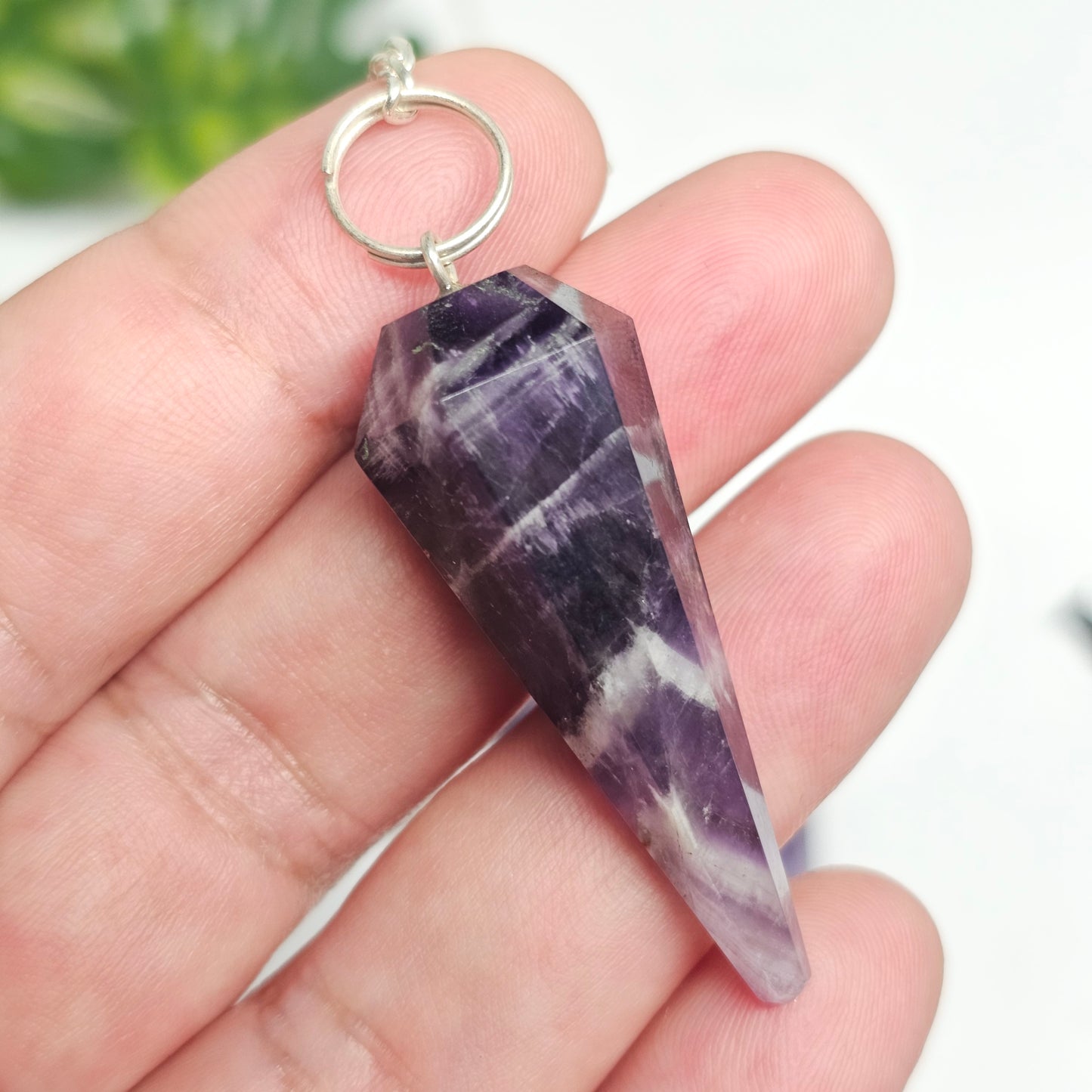 Amethyst pendulum held in hand to demonstrate its size, polished finish, crystal point, and chain for divination and energy work.