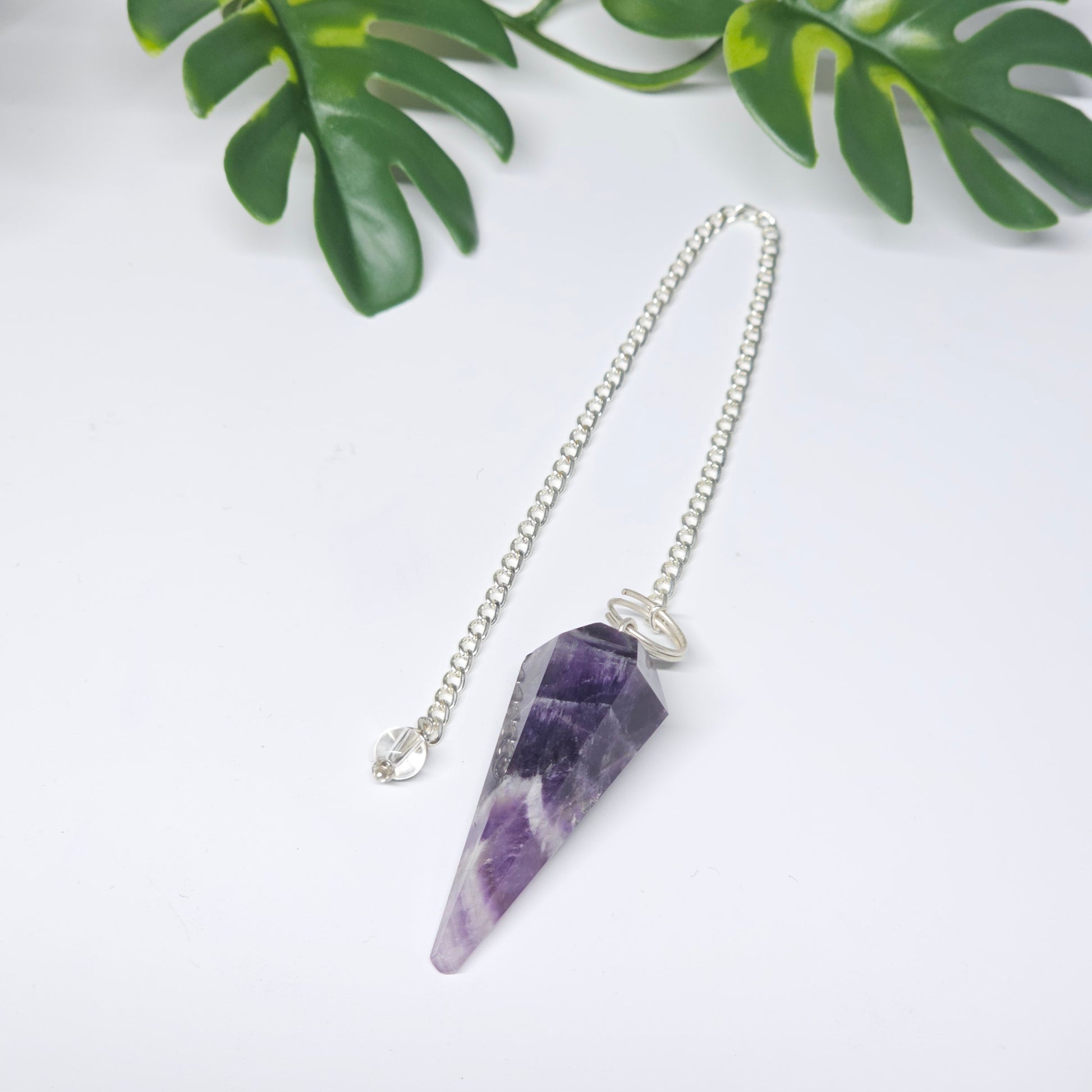 Close-up of an Amethyst pendulum featuring a polished crystal point, attached chain, and decorative bead, displayed on a flat surface.