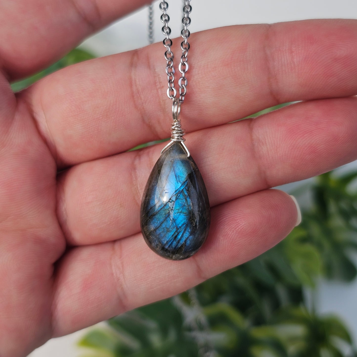 Handmade Labradorite pendant featuring striking blue flash and translucent depth. Wire wrapped in gold and suspended from a stainless steel chain, photographed across the hand and fingers in natural sunlight.