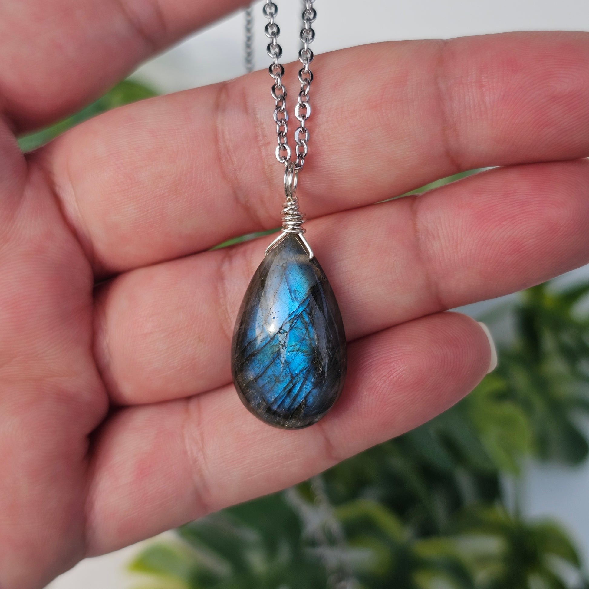Handmade Labradorite pendant featuring striking blue flash and translucent depth. Wire wrapped in gold and suspended from a stainless steel chain, photographed across the hand and fingers in natural sunlight.