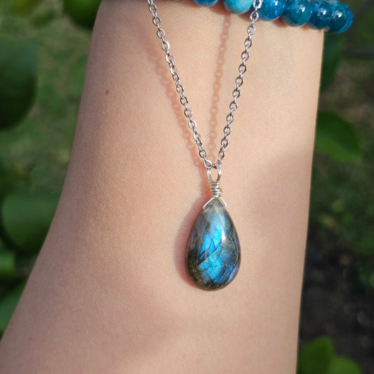Handmade Labradorite pendant with translucent high-flash blue iridescence, wire wrapped in gold and suspended from a stainless steel chain. Shown dangling from hand down the arm with sunlight enhancing its natural shimmer.
