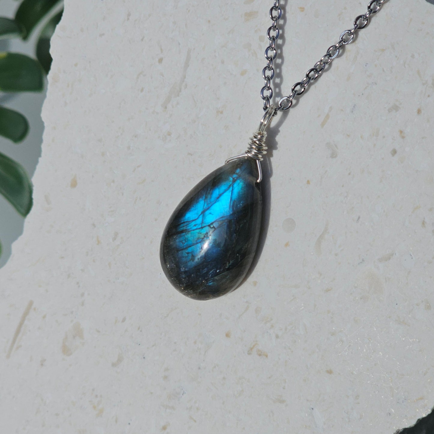 Handmade Labradorite pendant featuring translucent high-flash stone with striking blue iridescent flash. Wire wrapped in gold and suspended from a stainless steel chain, photographed hanging against a backdrop in natural sunlight.