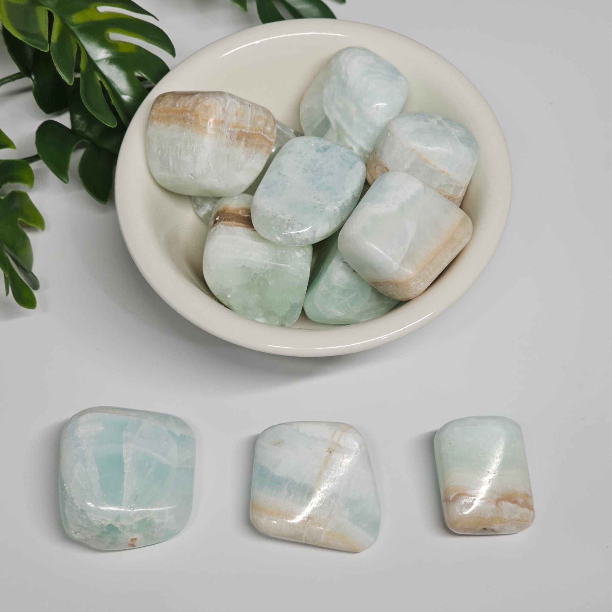 A collection of Caribbean Calcite tumble stones displayed in a bowl with one of each size shown separately on a flat surface, highlighting the difference between small, medium, and large pieces.