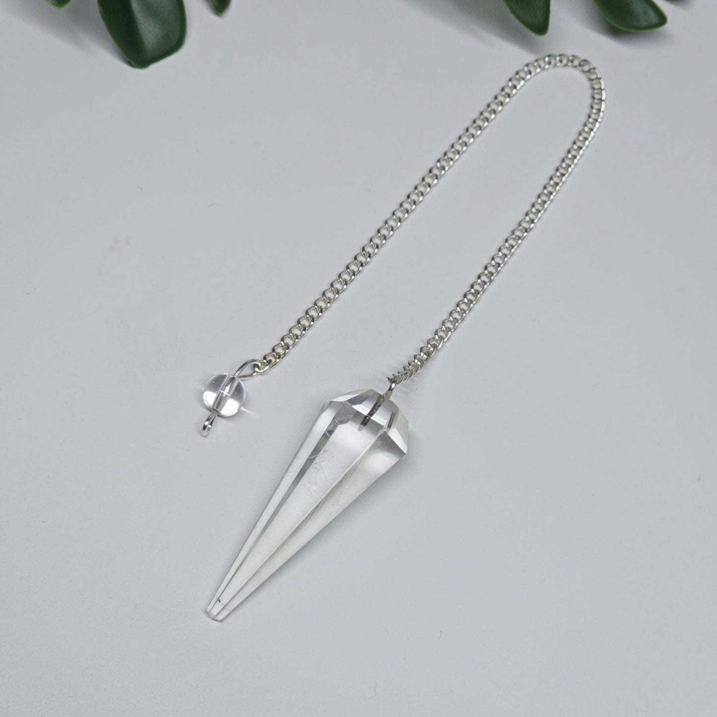Close-up of a Clear Quartz pendulum featuring a polished crystal point, attached chain, and decorative bead, displayed on a flat surface.