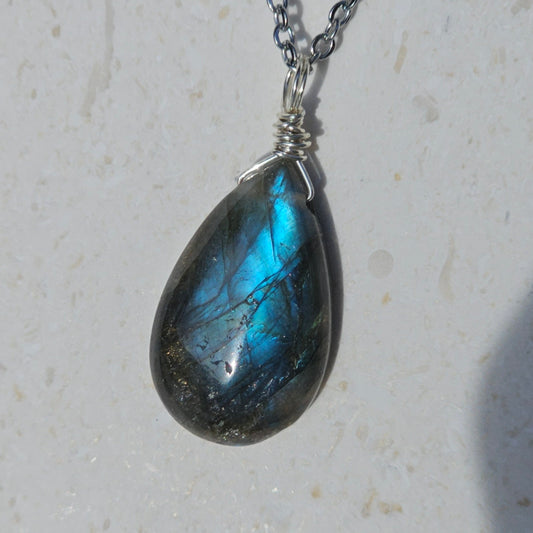 Close-up of a handmade Labradorite pendant featuring vivid blue iridescent flash and translucent depth. Wire wrapped in gold with a stainless steel chain, highlighted in natural sunlight.
