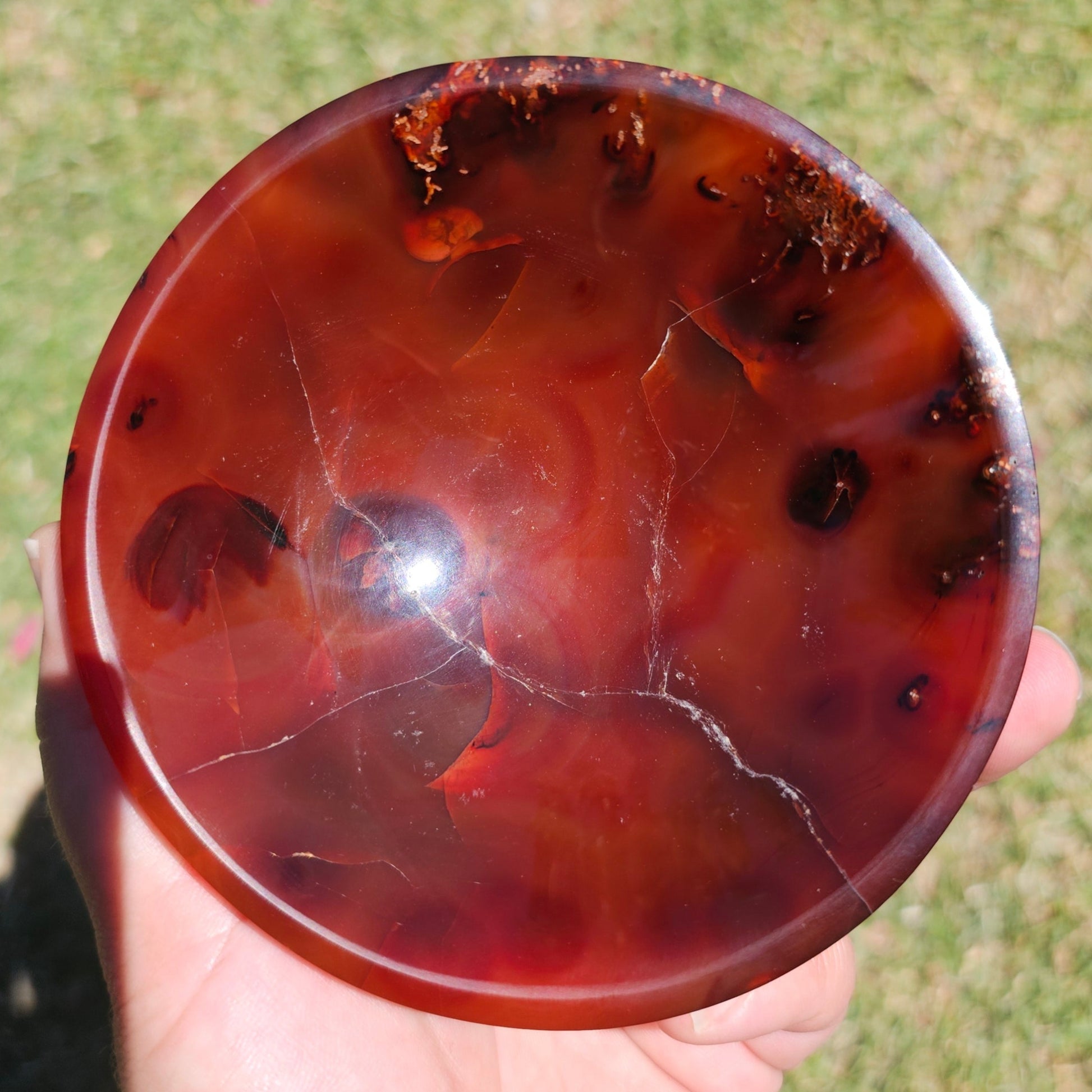 Top view of the dark Carnelian crystal bowl photographed in hand under natural sunlight, highlighting its rich warm tones, polished finish, and natural crystal patterns.