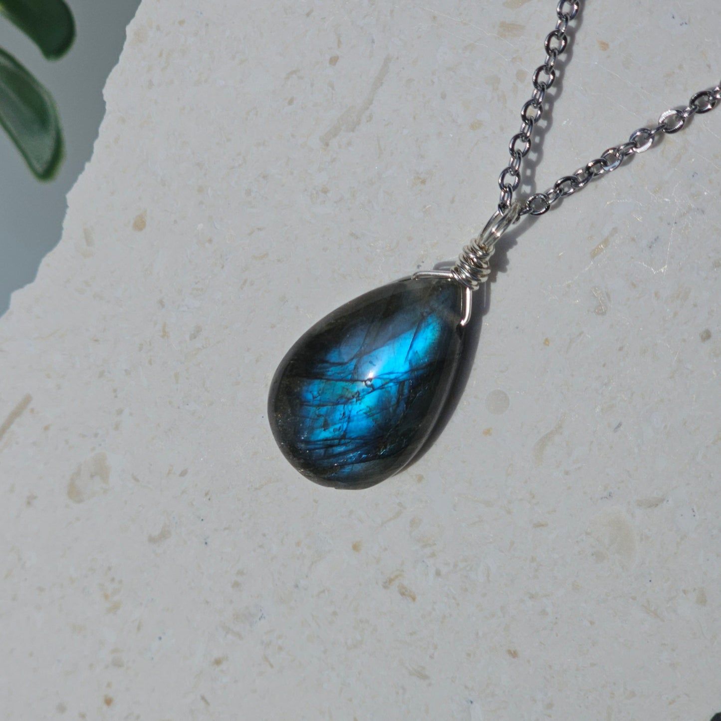 Handmade Labradorite pendant featuring translucent high-flash stone with striking blue iridescent flash. Wire wrapped in gold and suspended from a stainless steel chain, photographed hanging against a backdrop in natural sunlight.