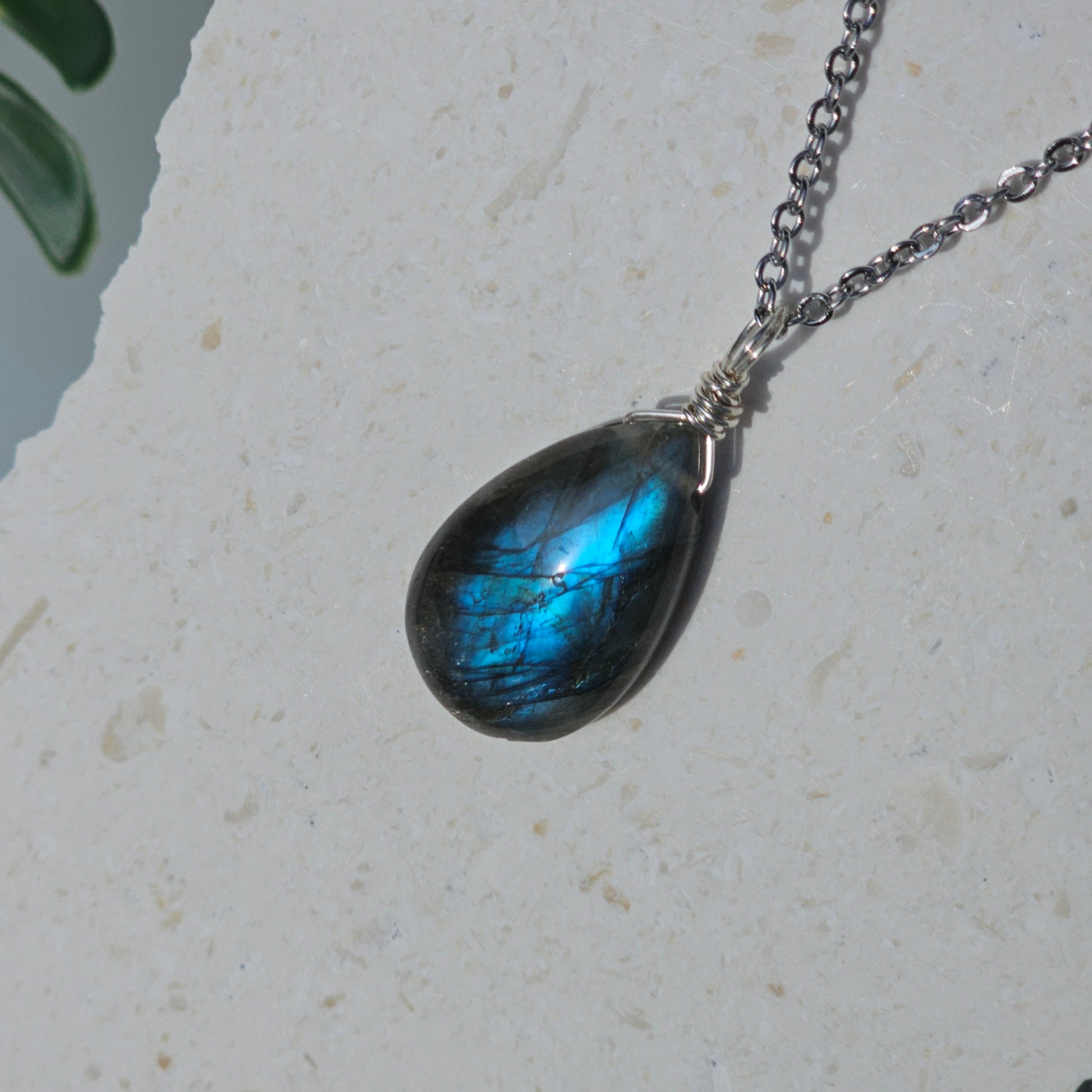 Handmade Labradorite pendant featuring translucent high-flash stone with striking blue iridescent flash. Wire wrapped in gold and suspended from a stainless steel chain, photographed hanging against a backdrop in natural sunlight.