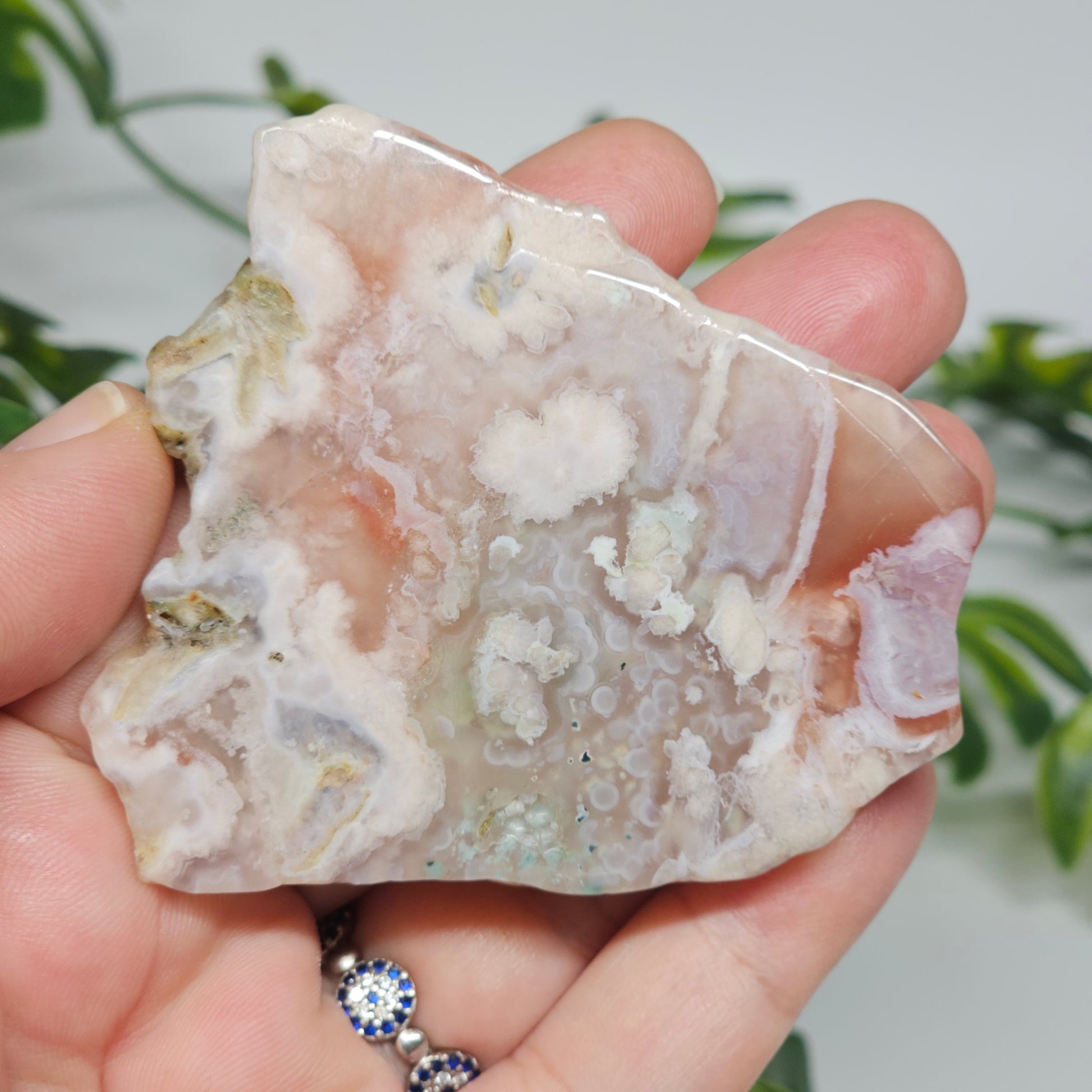 Polished Flower Agate slab held in hand to show natural size, warm pink-red tones, and floral plume inclusions. A nurturing crystal for growth, healing, and new beginnings.