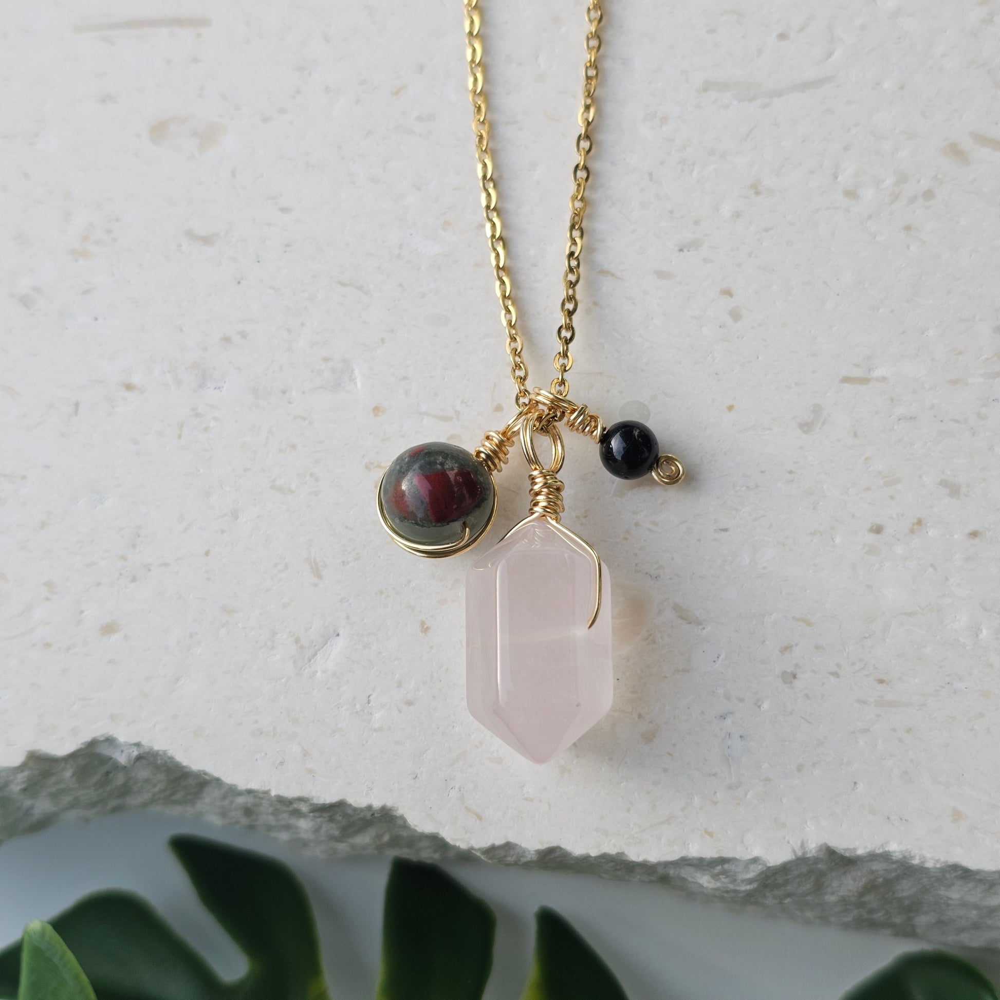 Flat lay of the Grounded Love Necklace showcasing a Rose Quartz point with Bloodstone Jasper and Black Tourmaline accents suspended from an 18k gold plated stainless steel cable chain