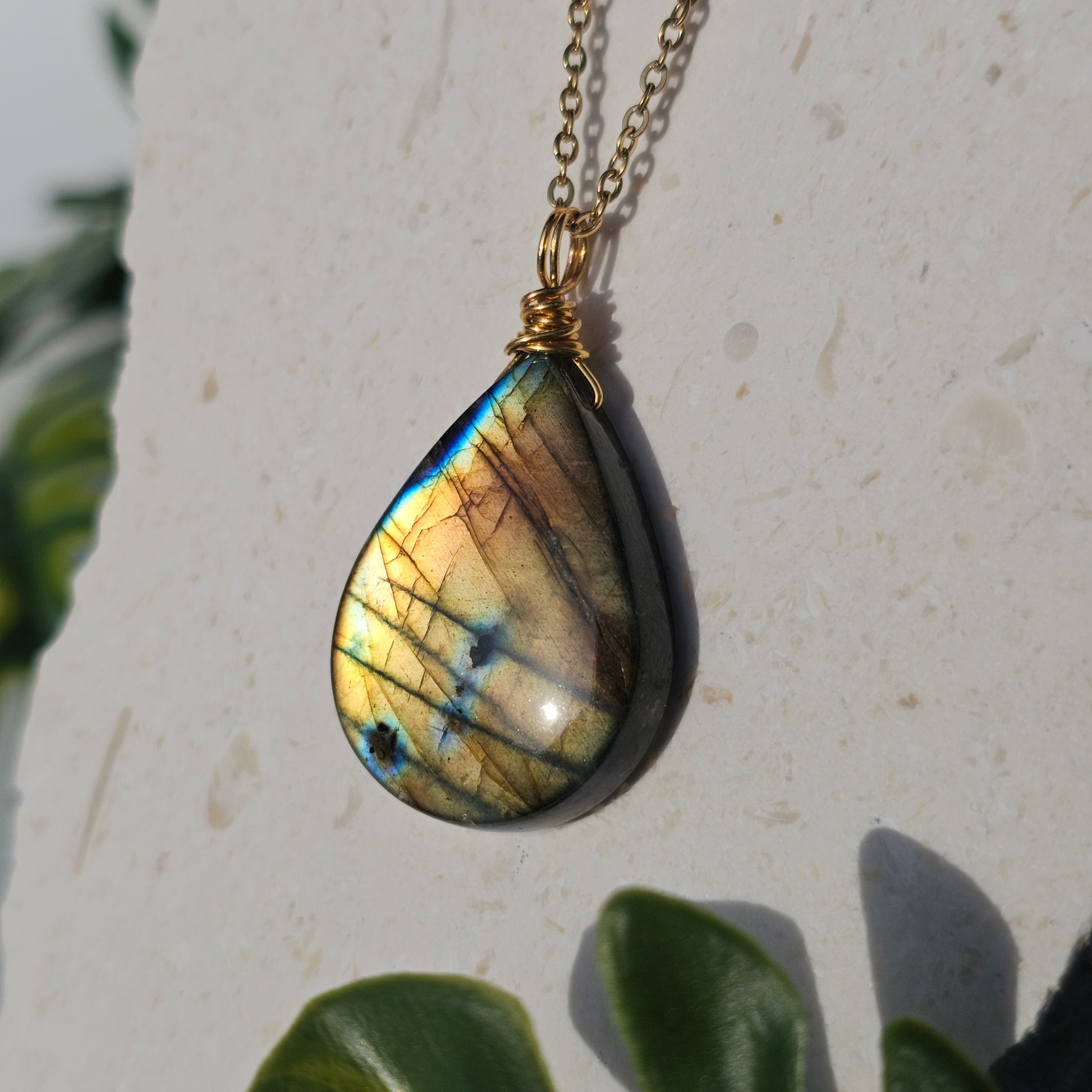 Handmade Labradorite pendant featuring high-flash stone with striking golden copper and blue iridescent flashes. Wire wrapped in gold and suspended from an 18k gold plated stainless steel chain, shown dangling down from backdrop in natural sunlight.
