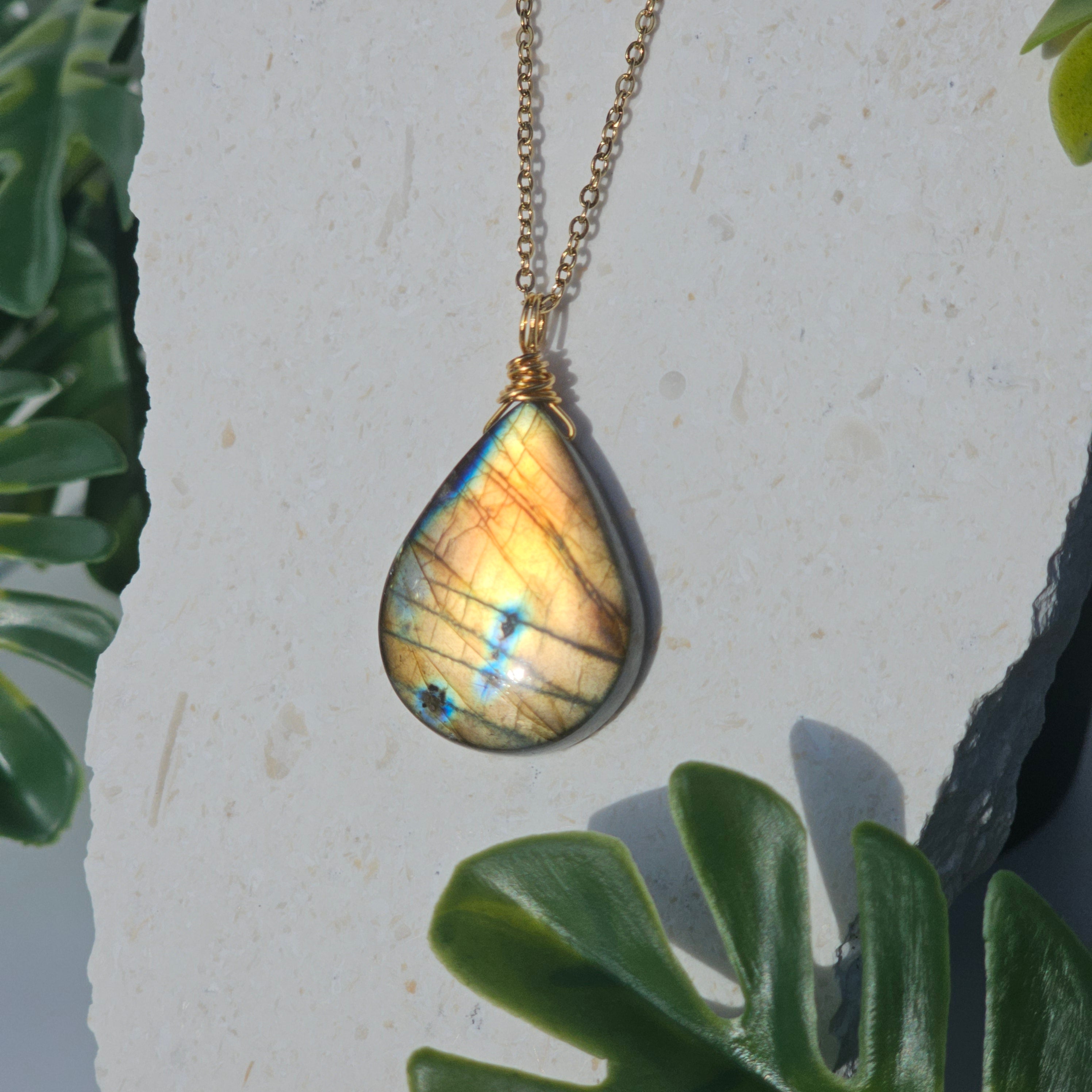 Handmade Labradorite pendant featuring high-flash stone with striking golden copper and blue iridescent flashes. Wire wrapped in gold and suspended from an 18k gold plated stainless steel chain, shown dangling from backdrop in natural sunlight.