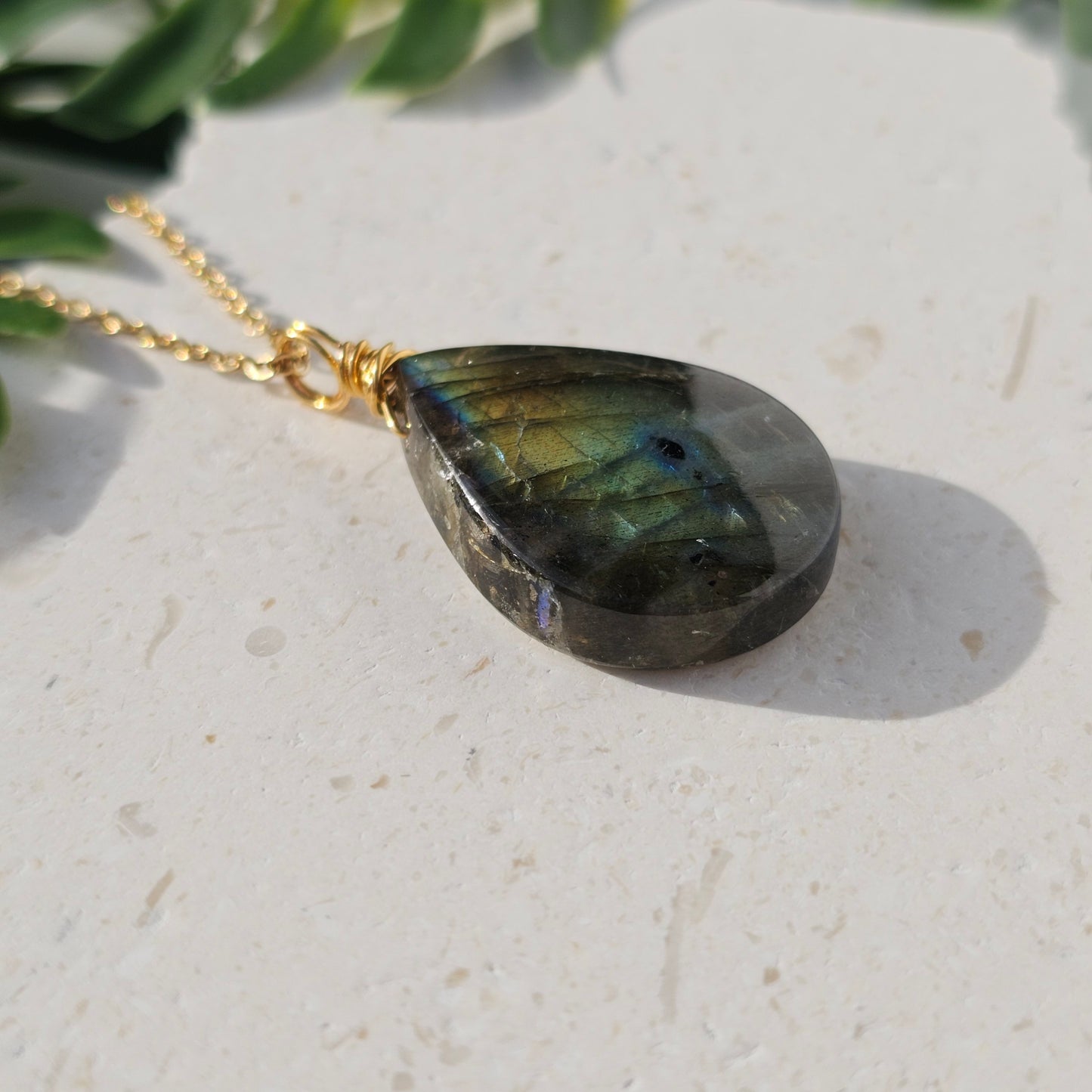 Handmade Labradorite pendant with high-flash stone displaying golden copper and blue iridescent flashes. Wire wrapped in gold and paired with an 18k gold plated stainless steel chain, shown flat from beneath with sunlight enhancing its natural shimmer.