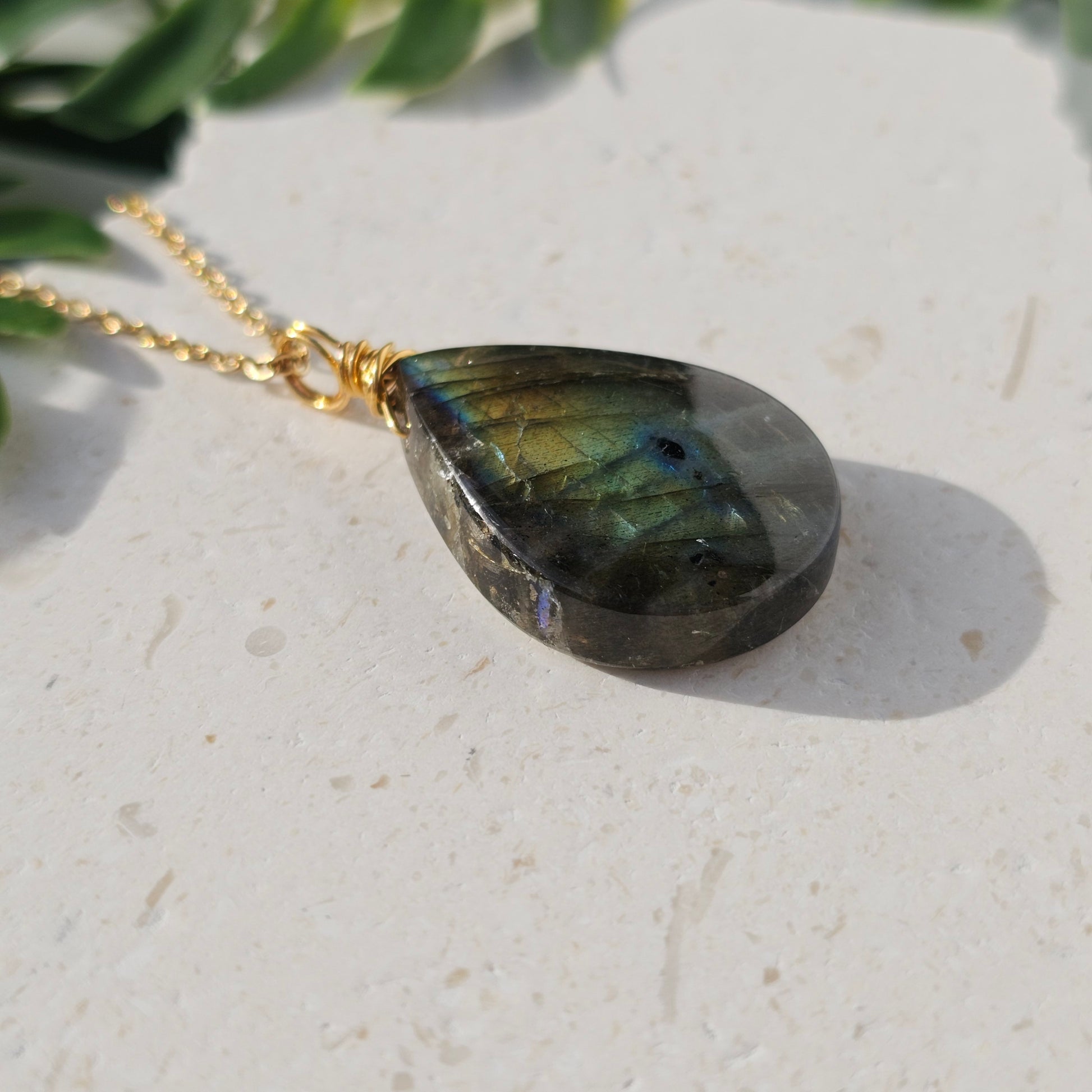 Handmade Labradorite pendant with high-flash stone displaying golden copper and blue iridescent flashes. Wire wrapped in gold and paired with an 18k gold plated stainless steel chain, shown flat from beneath with sunlight enhancing its natural shimmer.