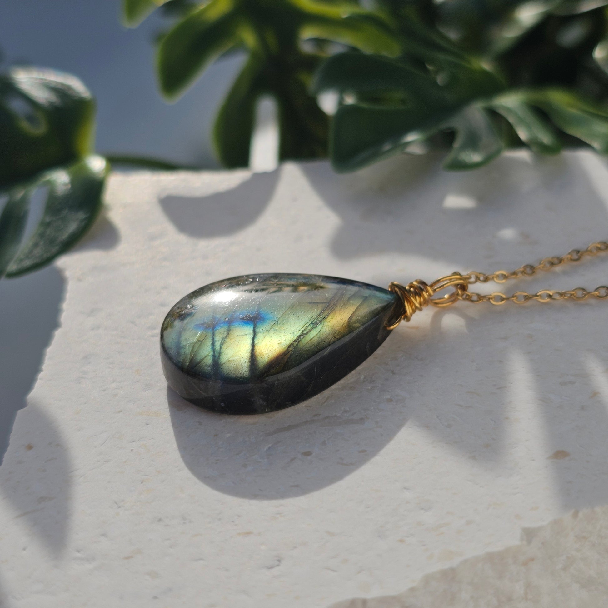 Handmade Labradorite pendant with high-flash stone displaying golden copper and blue iridescent flashes. Wire wrapped in gold and paired with an 18k gold plated stainless steel chain, shown flat with sunlight enhancing its natural shimmer.