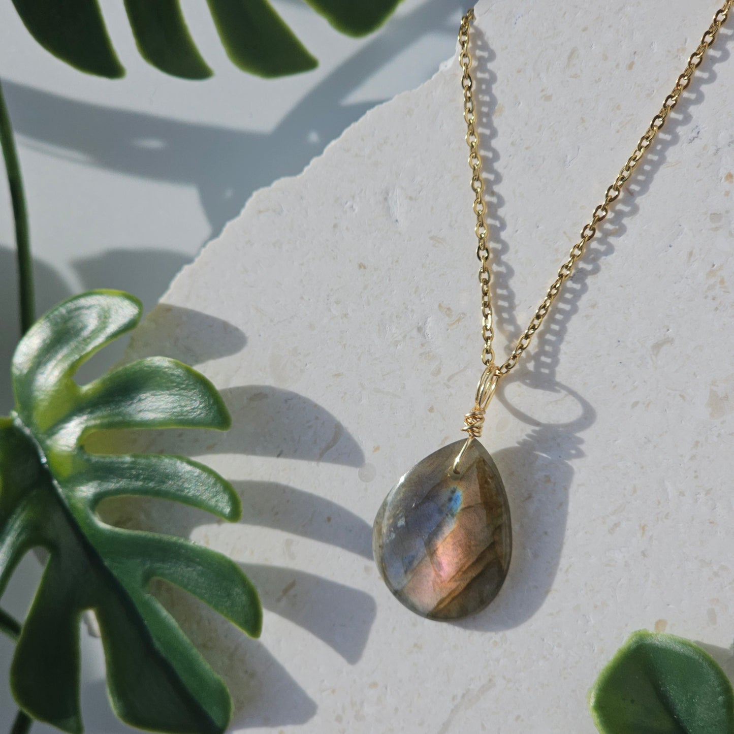Handmade Labradorite pendant with translucent high-flash stone showing pink and blue iridescent flashes. Wire wrapped in gold and paired with an 18k gold plated stainless steel chain, displayed flat with sunlight enhancing its natural shimmer.