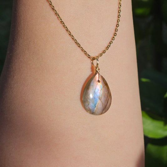 Handmade Labradorite pendant featuring a translucent high-flash stone with striking pink and blue iridescent flashes. Wire wrapped in gold and suspended from an 18k gold plated stainless steel chain, shown dangling from hand down the arm in natural sunlight.