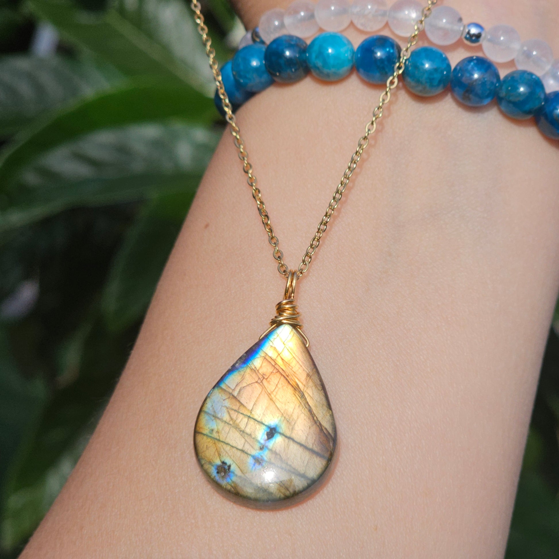 Handmade Labradorite pendant featuring high-flash stone with striking golden copper and blue iridescent flashes. Wire wrapped in gold and suspended from an 18k gold plated stainless steel chain, shown dangling from hand down the arm in natural sunlight.