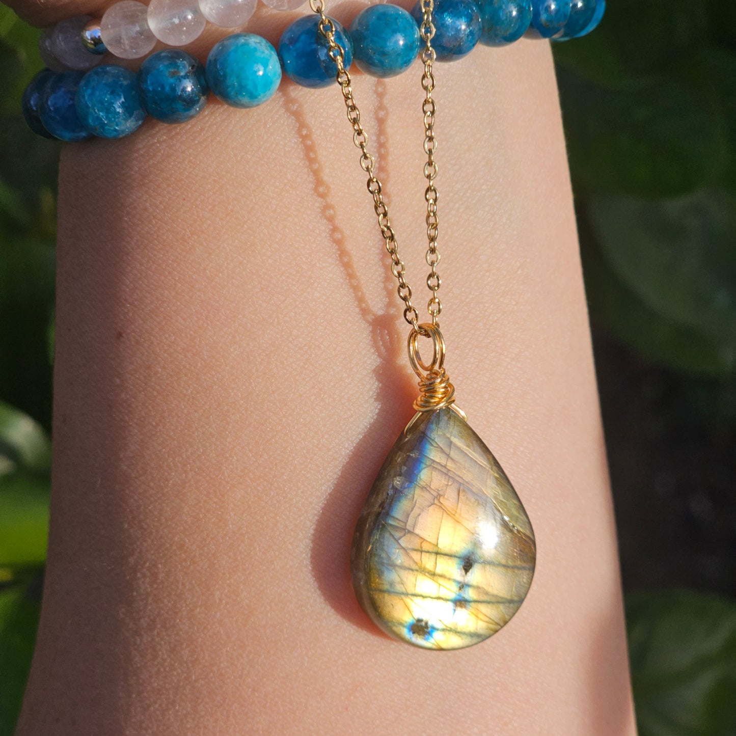 Handmade Labradorite pendant featuring high-flash stone with striking golden copper and blue iridescent flashes. Wire wrapped in gold and suspended from an 18k gold plated stainless steel chain, shown dangling from hand down the arm in natural sunlight.