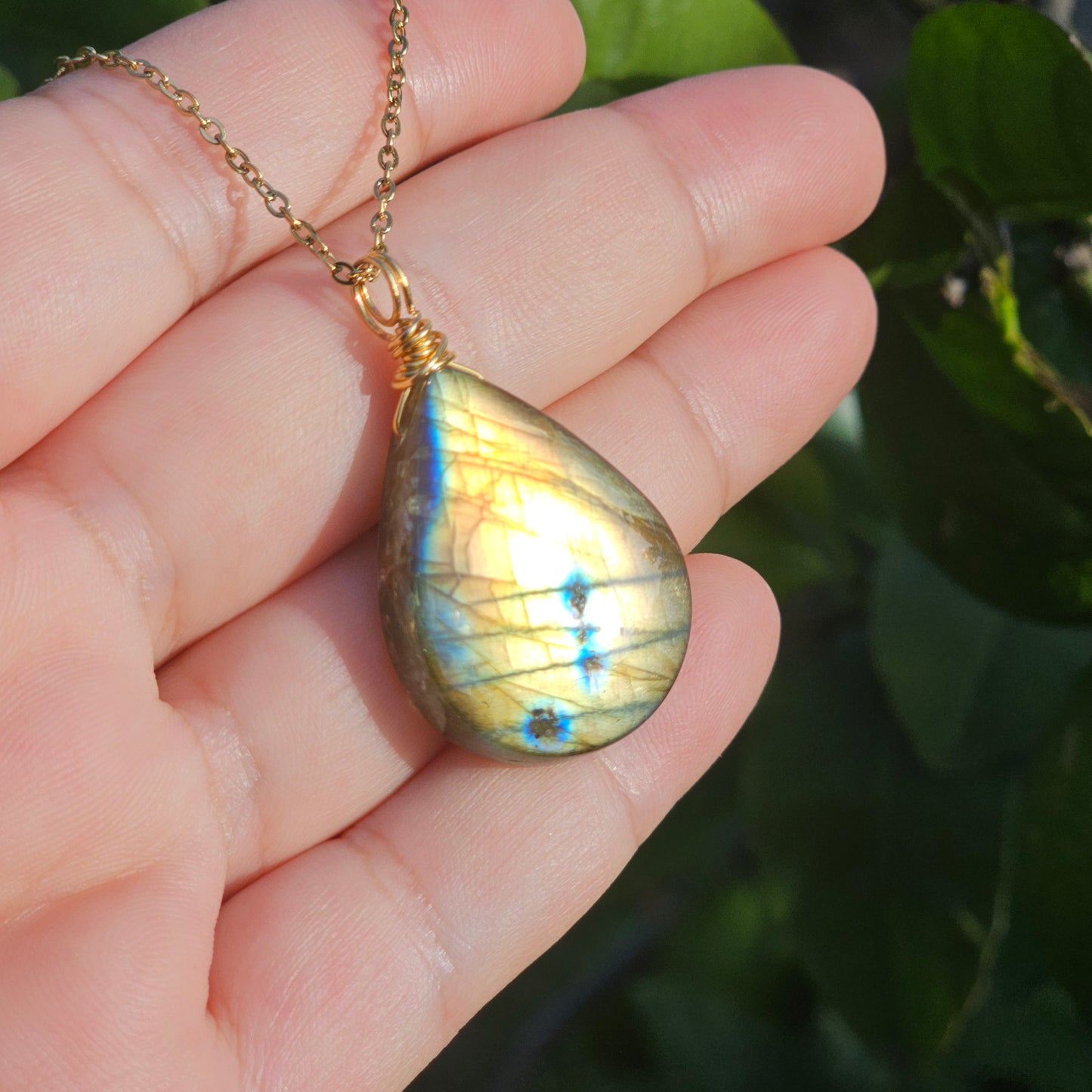 Handmade Labradorite pendant featuring high-flash stone with striking golden copper and blue iridescent flashes. Wire wrapped in gold and suspended from an 18k gold plated stainless steel chain, shown on hand in natural sunlight.