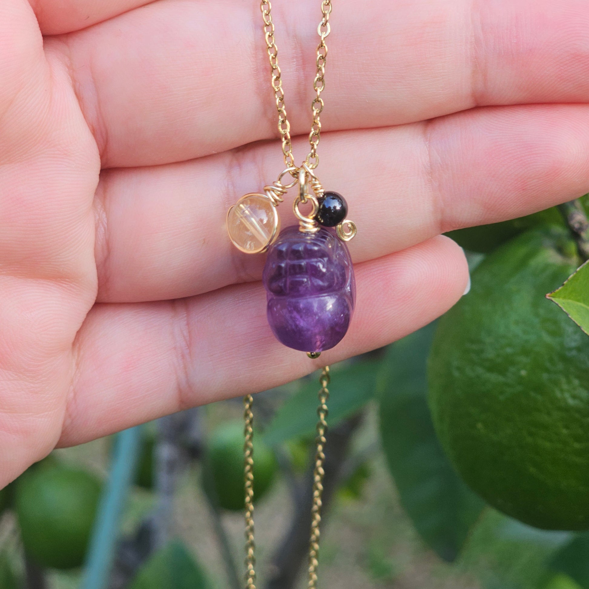 Close up of the Protected Prosperity Necklace hanging from a hand highlighting Amethyst Pixiu with Citrine and Black Tourmaline gemstone beads