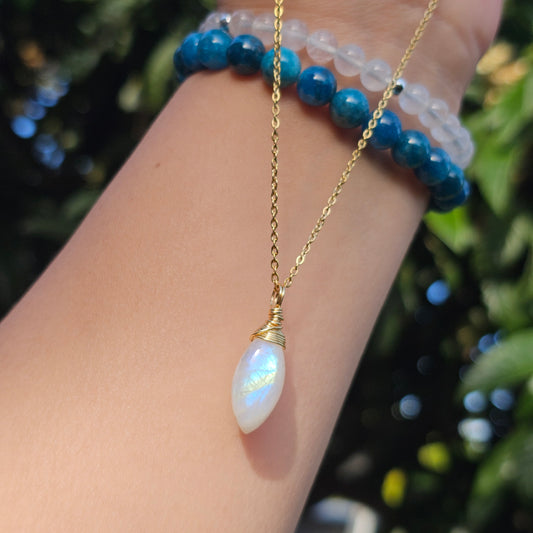 Rainbow Moonstone marquise cabochon pendant with golden blue flash, wire wrapped in gold and suspended from a stainless steel chain. Photographed dangling from hand down the arm with sunlight enhancing its translucent glow and natural iridescence.