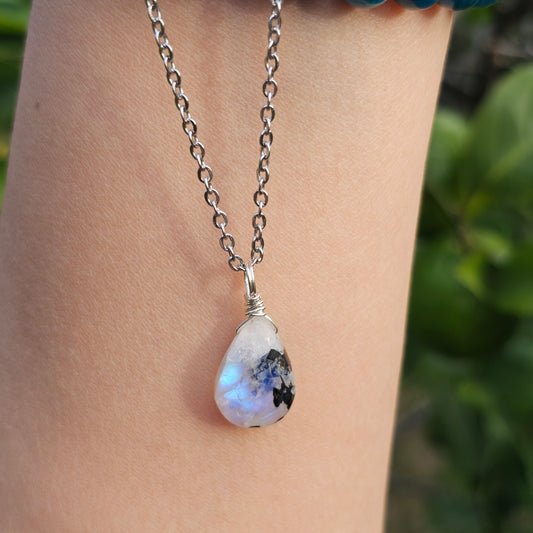 Handmade Rainbow Moonstone pendant featuring natural black tourmaline inclusions and ethereal blue flash. Wire wrapped in gold and suspended from a stainless steel chain, shown dangling from hand down the arm in natural sunlight.