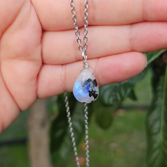 Handmade Rainbow Moonstone pendant featuring natural black tourmaline inclusions and ethereal blue flash. Wire wrapped in gold and suspended from a stainless steel chain, photographed hanging across the hand and fingers with sunlight enhancing its natural glow.