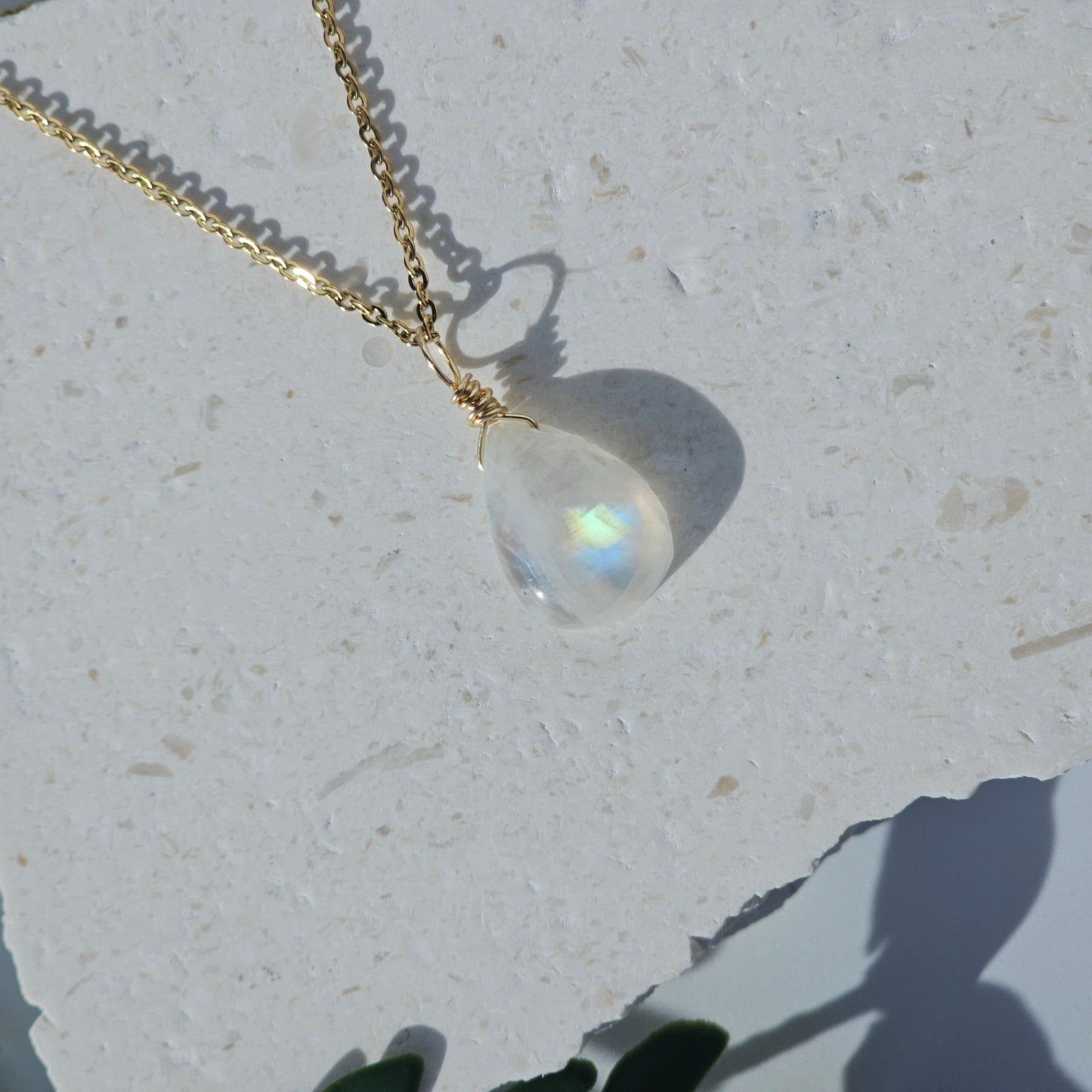 Rainbow Moonstone teardrop necklace featuring translucent stone with golden blue flash, wrapped in gold wire and paired with a stainless steel chain. Shown flat with sunlight highlighting its natural iridescence and glow.