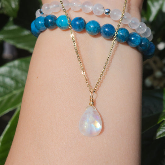 Rainbow Moonstone teardrop necklace with golden blue flash, wire wrapped in gold and suspended from a stainless steel chain. Photographed dangling from hand down the arm with sunlight enhancing its natural shimmer and translucency.